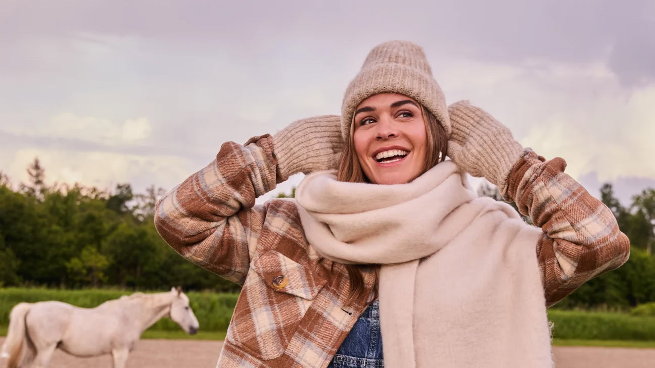 Femme souriante portant un manteau à carreaux brun, une tuque en tricot et un grand foulard beige à franges, dans une ambiance automnale à la campagne.